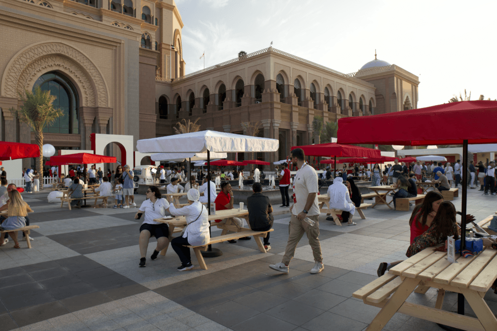 A bustling outdoor dining area with red umbrellas, wooden tables, and people enjoying their time, showcasing image.