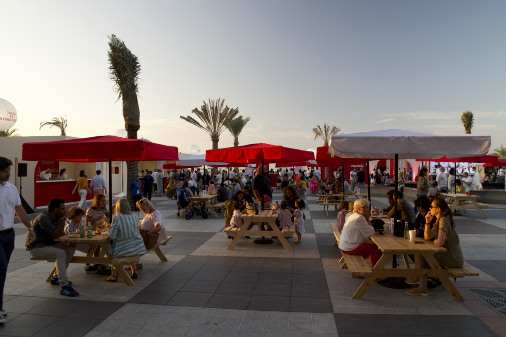 A bustling outdoor dining area with red umbrellas, people enjoying meals, and palm trees, showcasing image.
